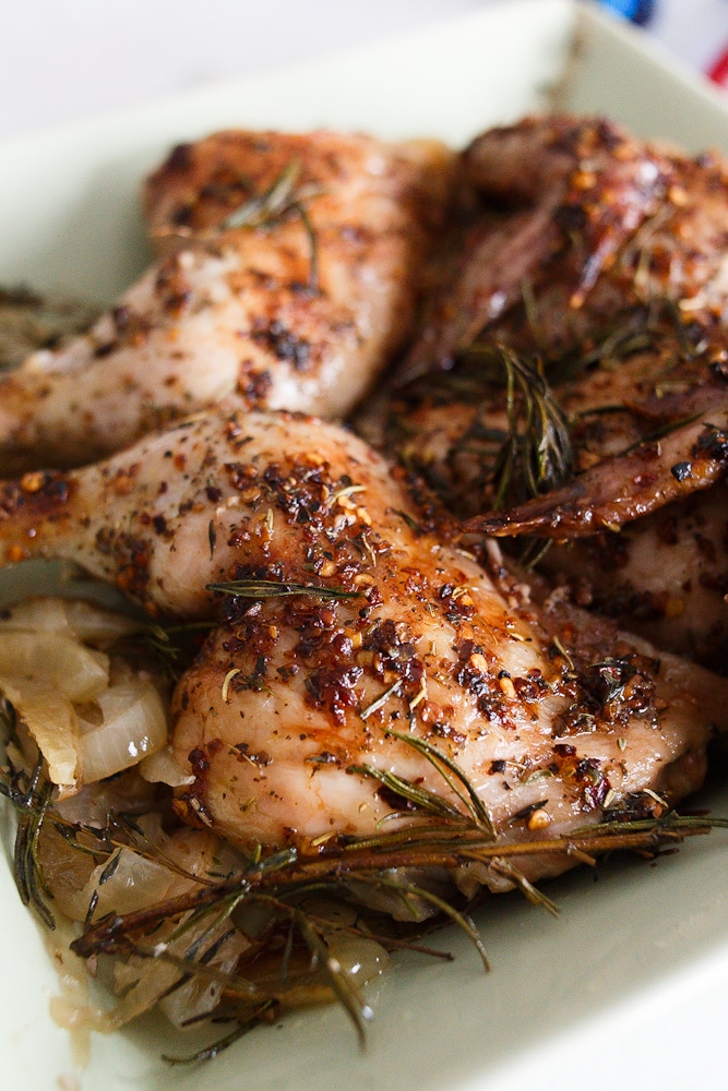 In a green casserole dish sits a chicken with both breasts and legs out to each side, cooked and topped with rosemary and thyme. 